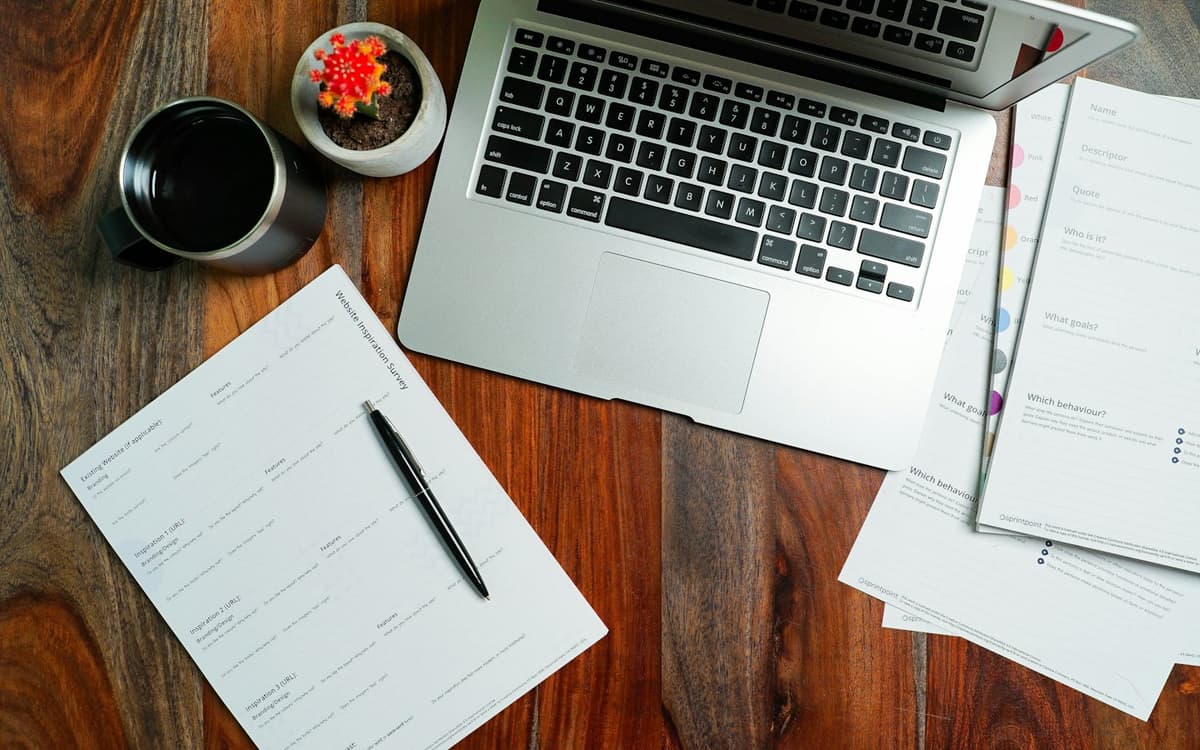 Printed forms and a pen beside a laptop on a wooden desk