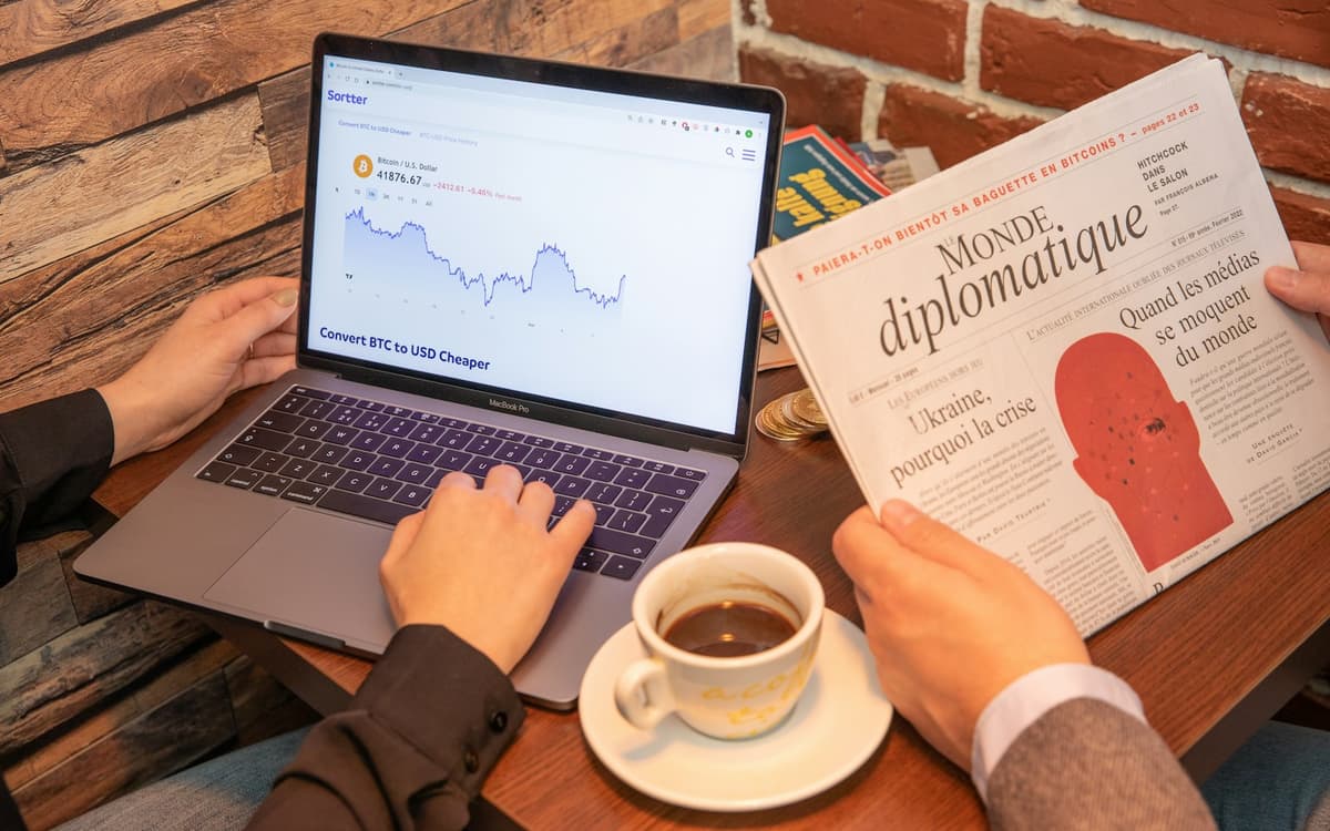 A person reading a newspaper beside a laptop and a cup of coffee